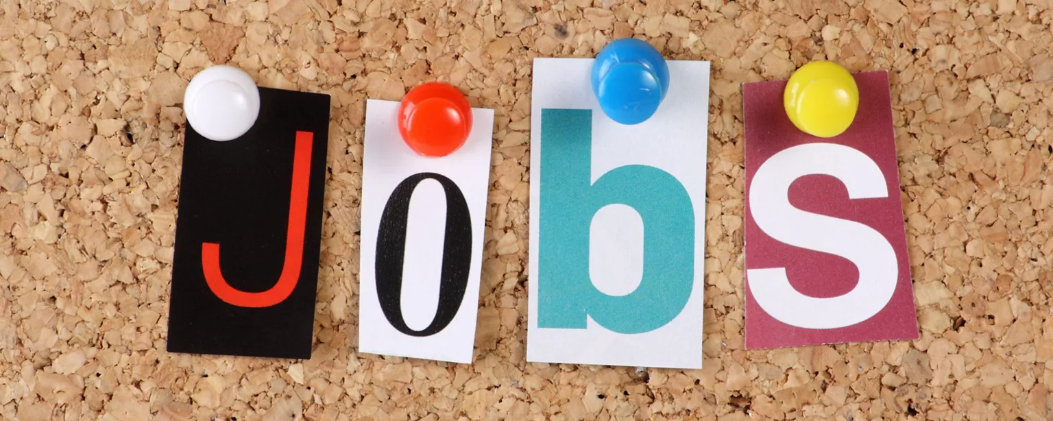 A cork bulletin board with four sticky notes of different colors pinned to it; the sticky notes spell out the word “JOBS.”