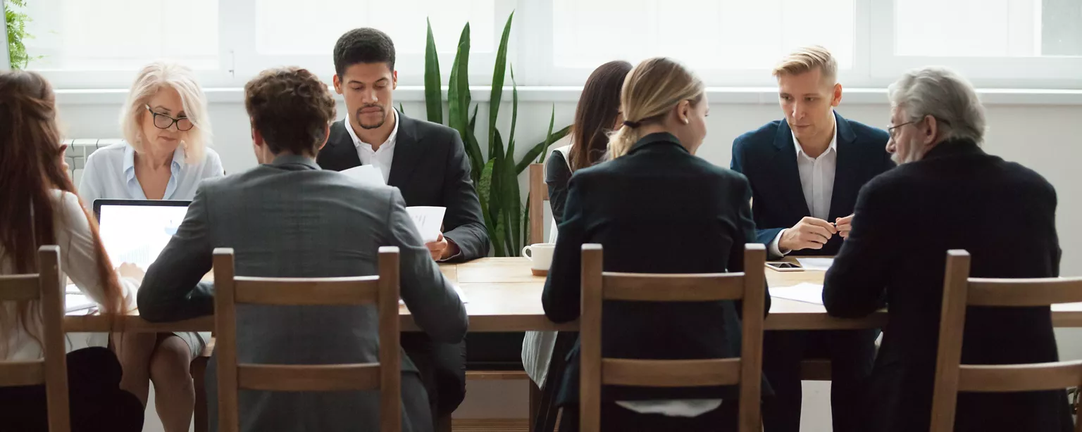 A group of multicultural leaders sit at a long table and discuss succession planning.