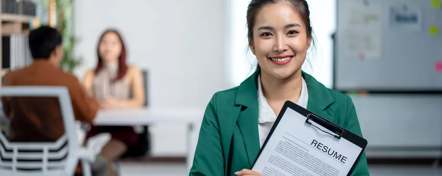 A woman with a resume attached to a clipboard stands and smiles as two people converse at a table in the background.