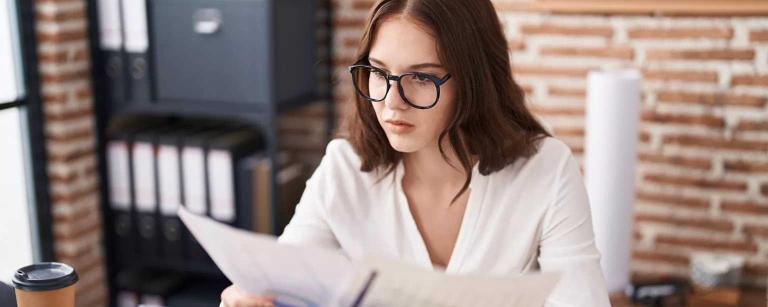 A focused young professional in glasses reviews 401(k) documents at her desk with a cup of coffee.