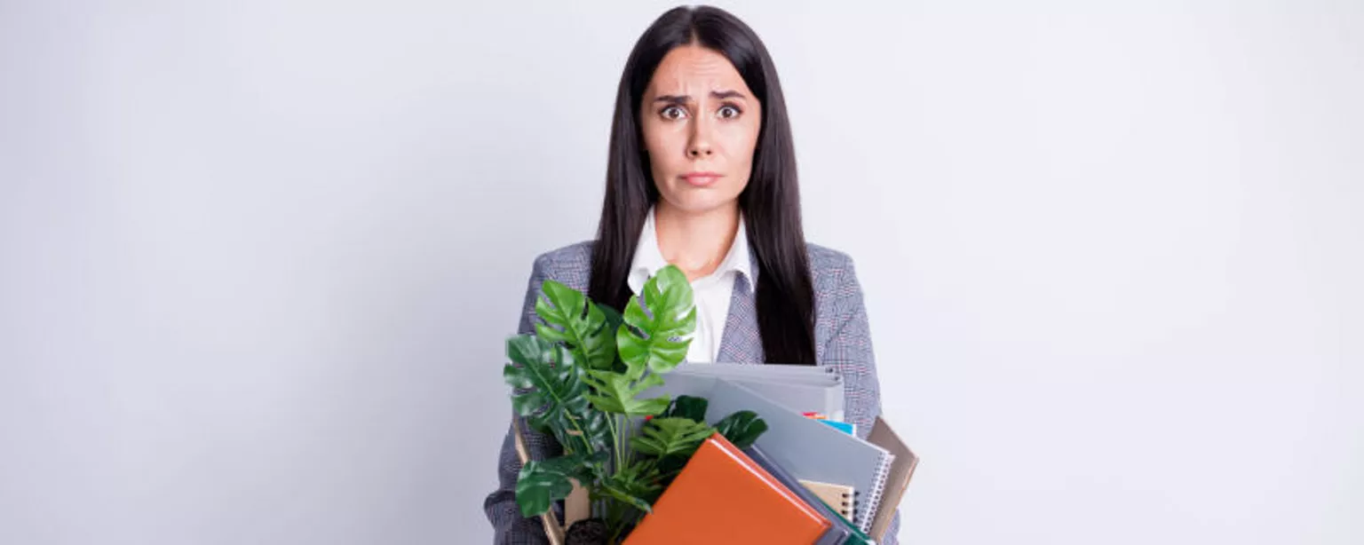 A woman carrying a small box with her belongings looks stunned as the reality of being laid off soaks in.