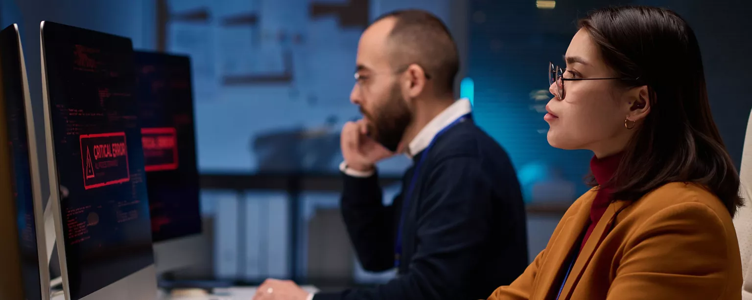 Two cybersecurity professionals sit at their workstations responding to a cyberattack; their computer screens show a warning: Critical Error.