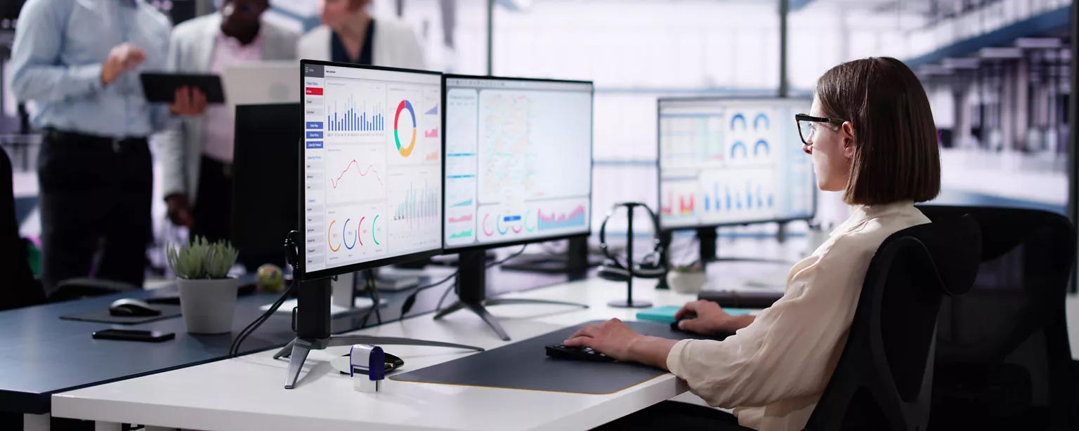 A data analyst in a corporate accounting department looks at financial charts and data appearing on three computer screens.