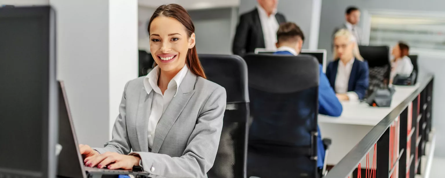 A smiling professional in a gray suit works on a laptop at her desk in a busy office.