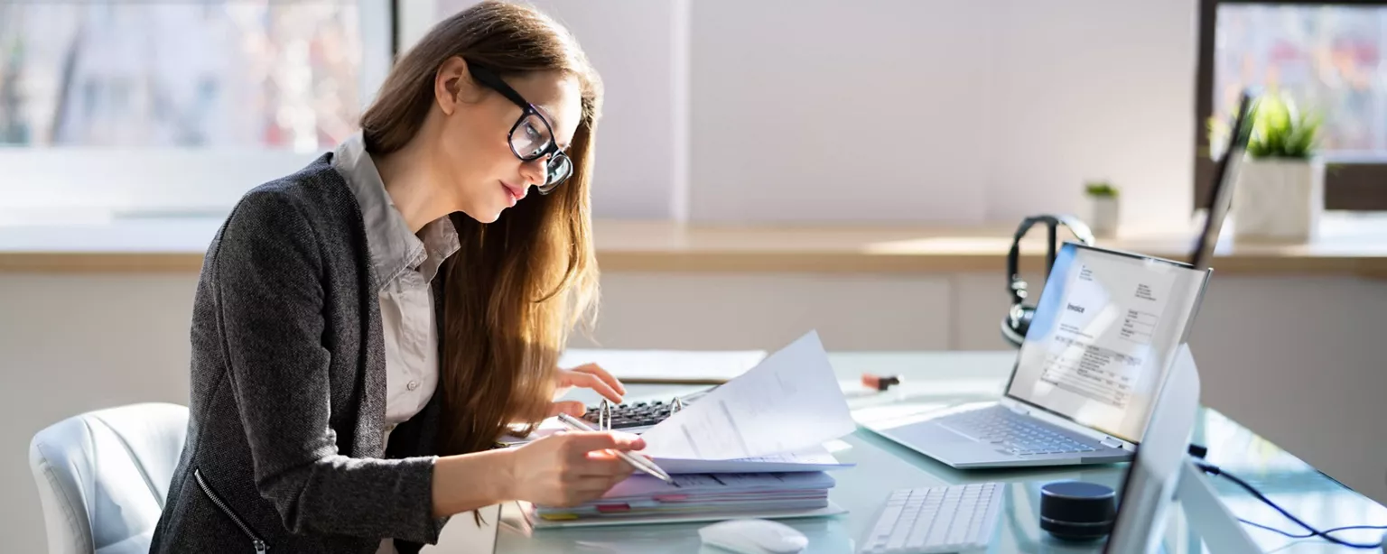 A public accountant reviews financial documents in a binder. On the laptop in front of her, the screen shows a record of an invoice.