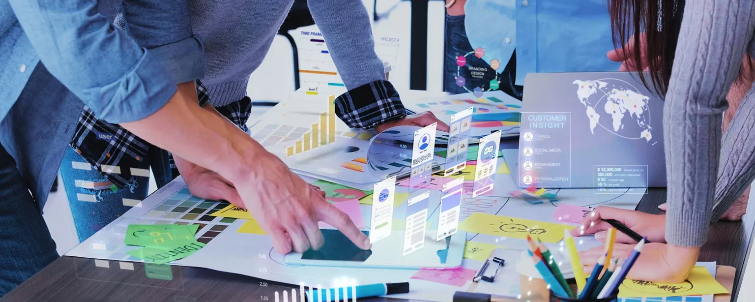 The hands of marketing and creative professionals are shown as they brainstorm as a team at a table in the office, with digital iconography superimposed.