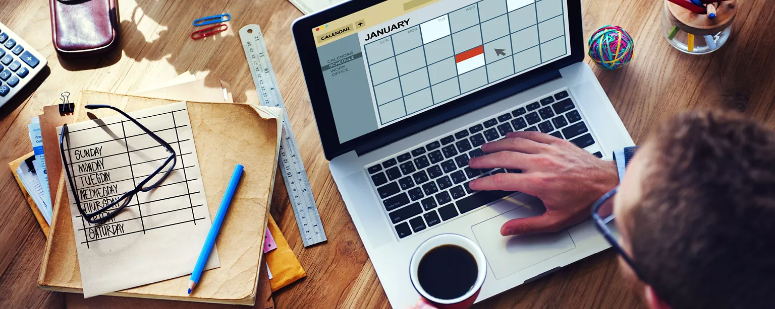 A person using a laptop to check a digital calendar while holding a coffee mug, surrounded by office supplies on a wooden desk.