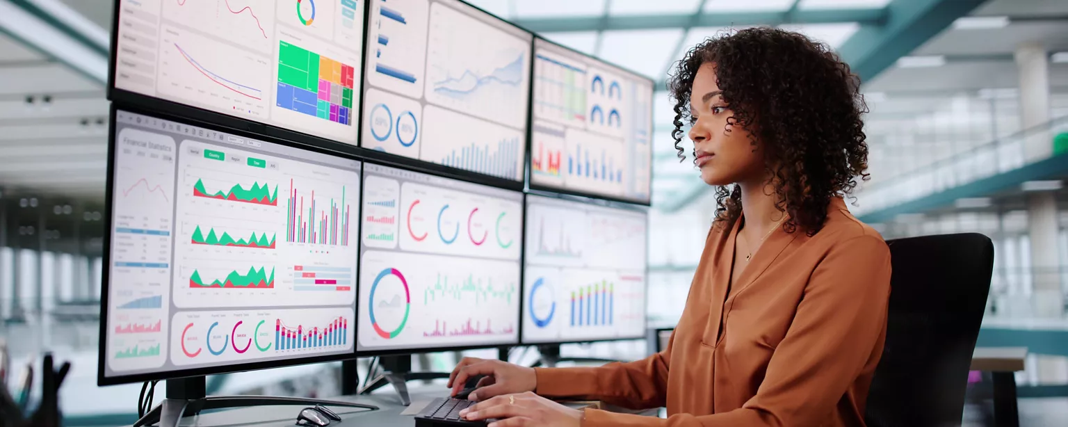 A financial analyst working at a desk reviews multiple large monitors displaying charts, graphs and financial data dashboards.