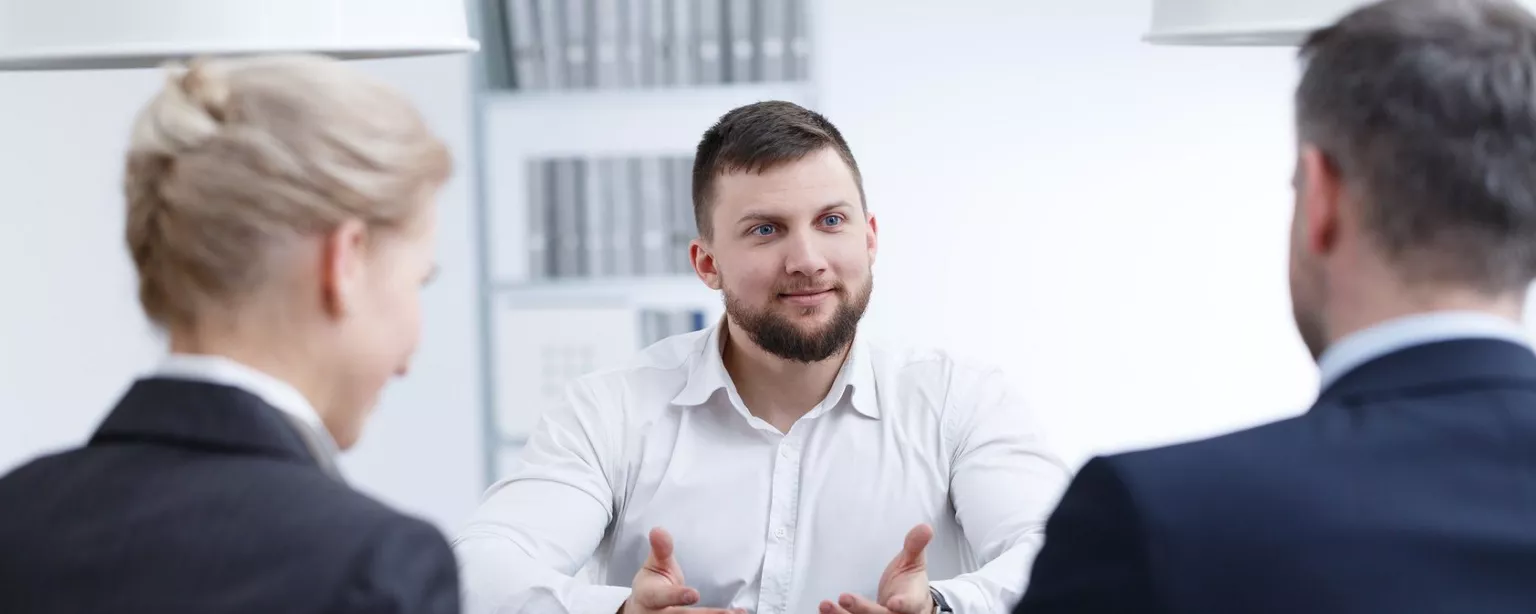 Junger Mann sitzt zwei formal gekleideten Gesprächspartnern gegenüber