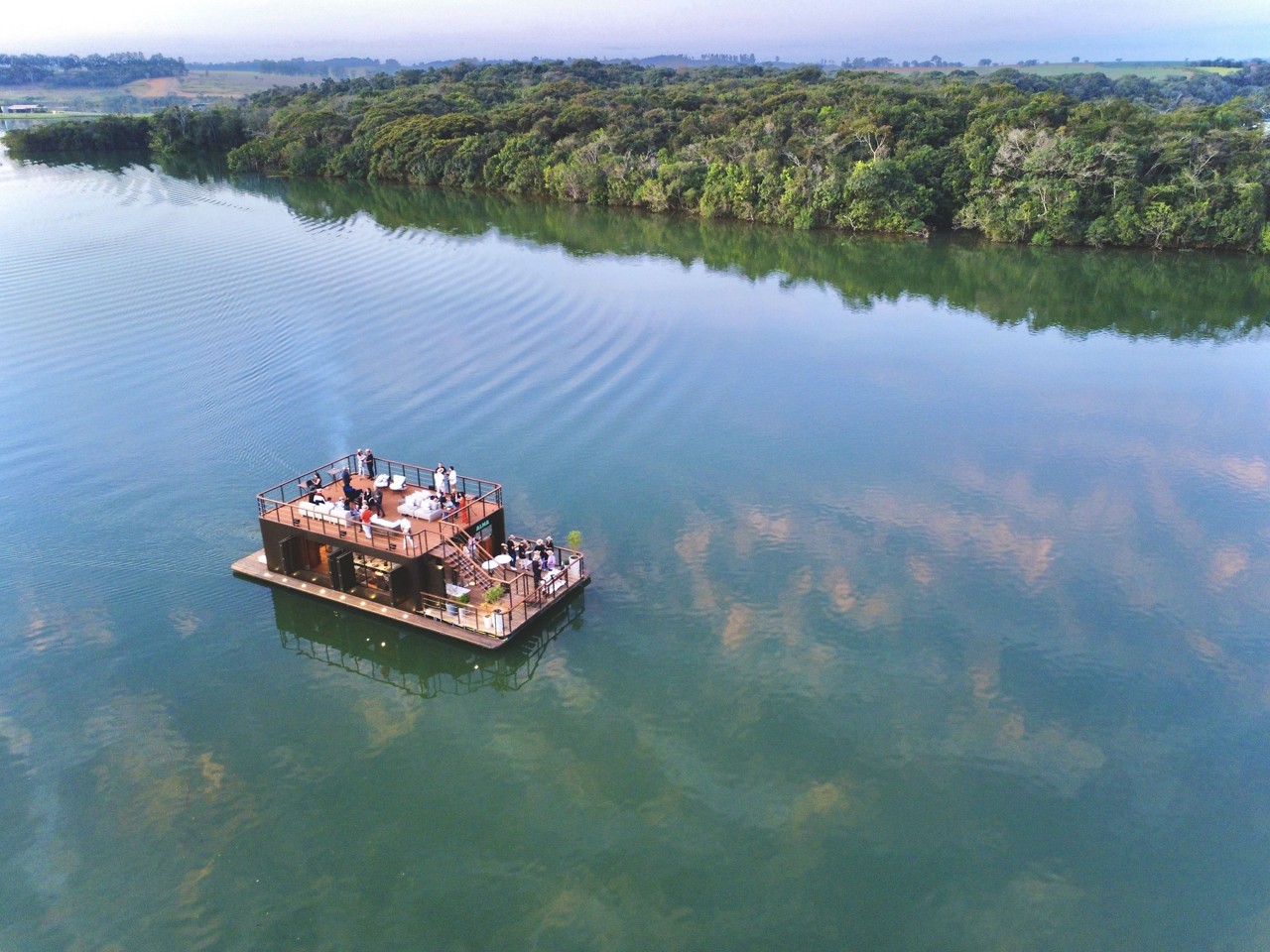 O wine bar flutuante navega pela represa do Funil, tendo ao fundo o complexo Alma Gerais