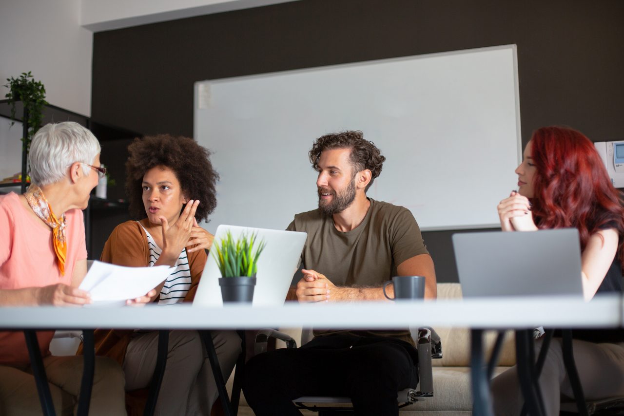 A group of four employees in a meeting room. Two of them have their laptops. One is holding some papers. The fourth is listening with his hands clasped.