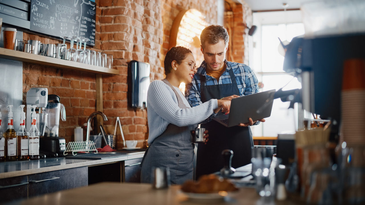 two people in aprons in a cafe reviewing documents on a laptop
