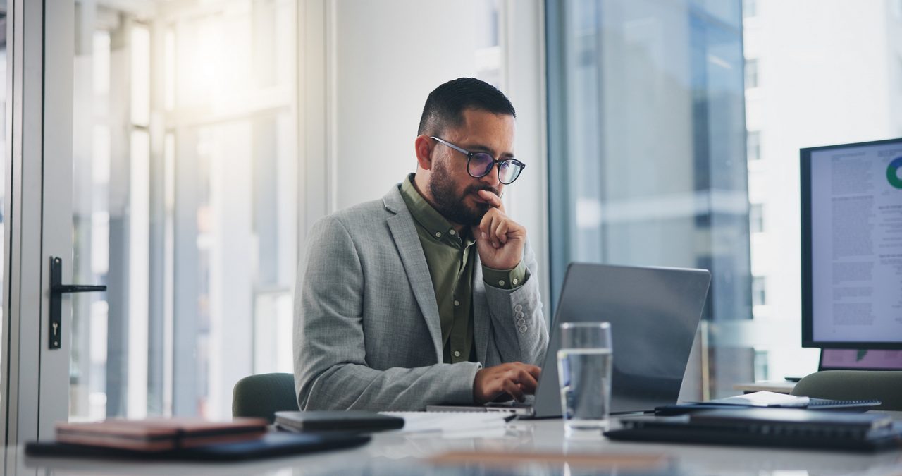 man with glasses sitting at desk looking carefully at a laptop screen