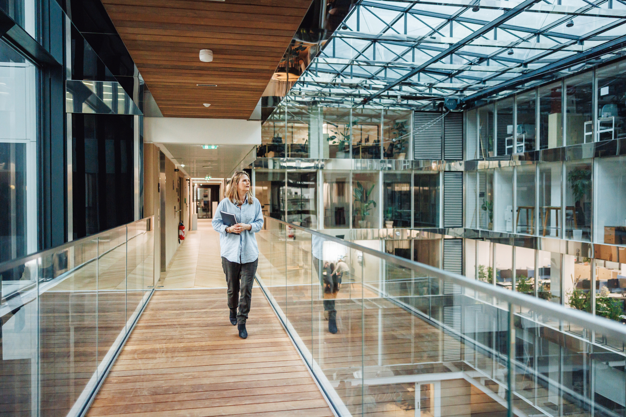 A mature Caucasian woman in business attire confidently holds a file folder while walking through the glass-walled hallway of a contemporary office.