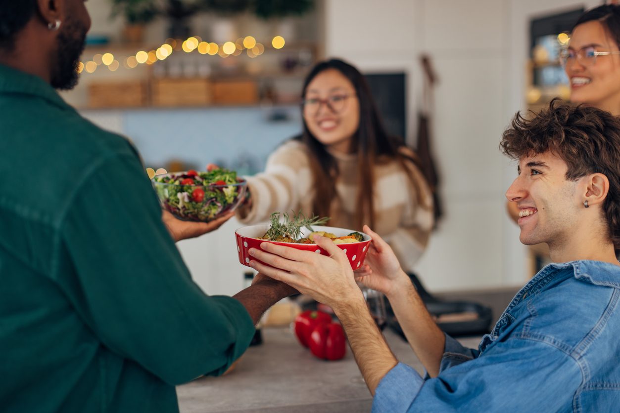 A Black man brings dishes of food to the table where his friends smile and help serve the food