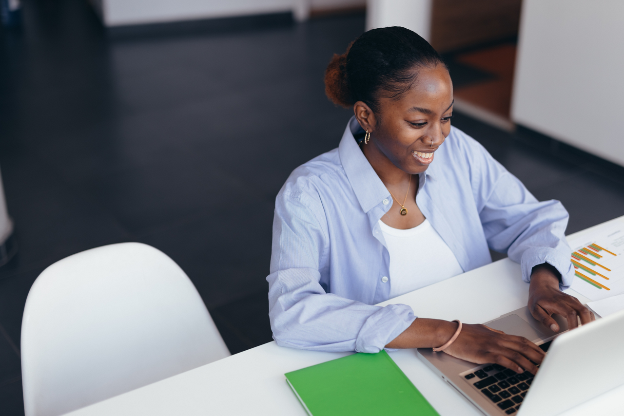 Young African American businesswoman smiling while working on her laptop, sitting at a white table with a green notebook and business charts