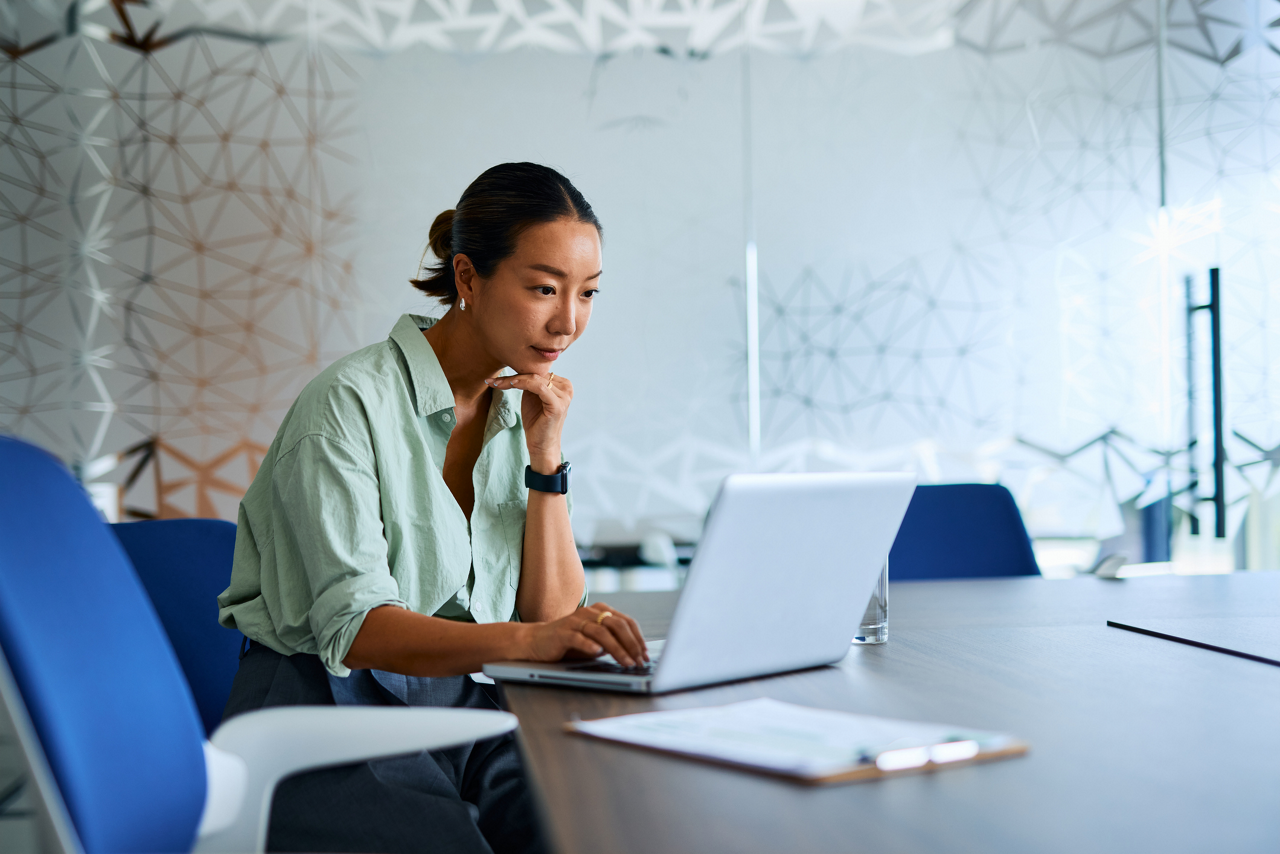 Asian businesswoman sitting at a meeting table, working intently on her laptop while resting her hand on her chin, deeply concentrating on her tasks and problem-solving strategies