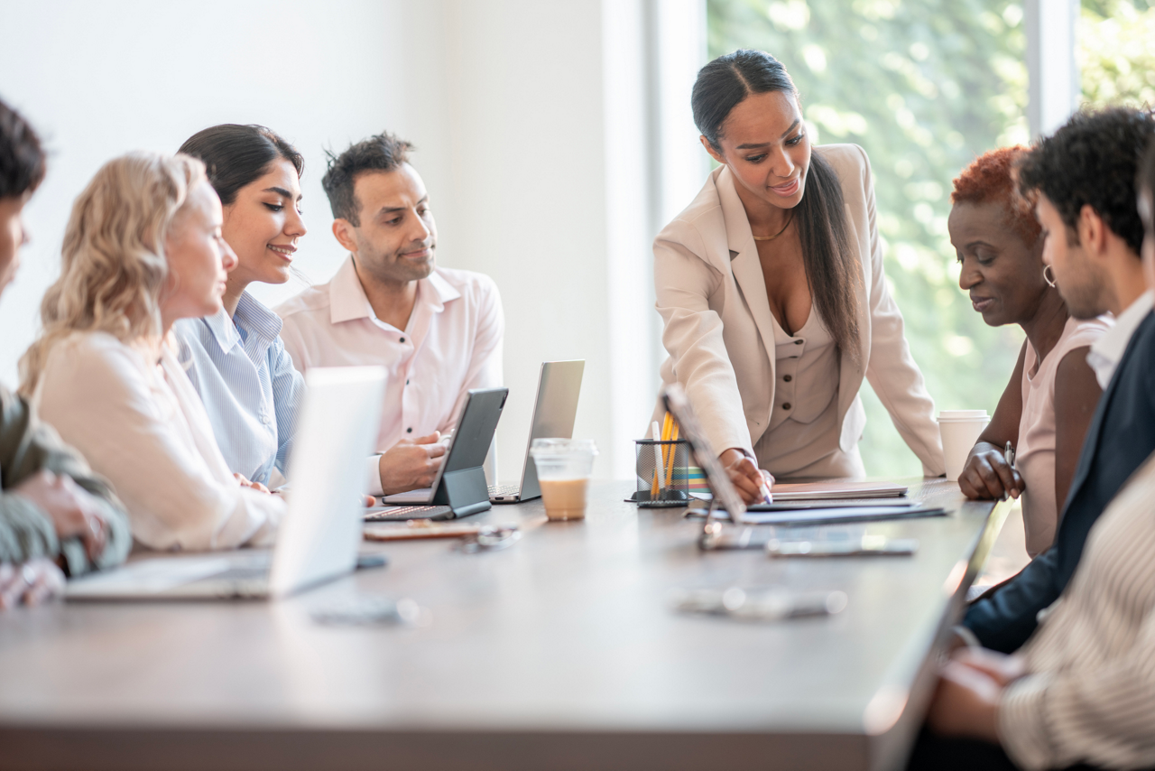 Business professionals engaged in a collaborative discussion around a conference table in a bright office space. The team emphasizes collaboration, diversity, and teamwork in their pursuit of shared goals and innovative ideas.