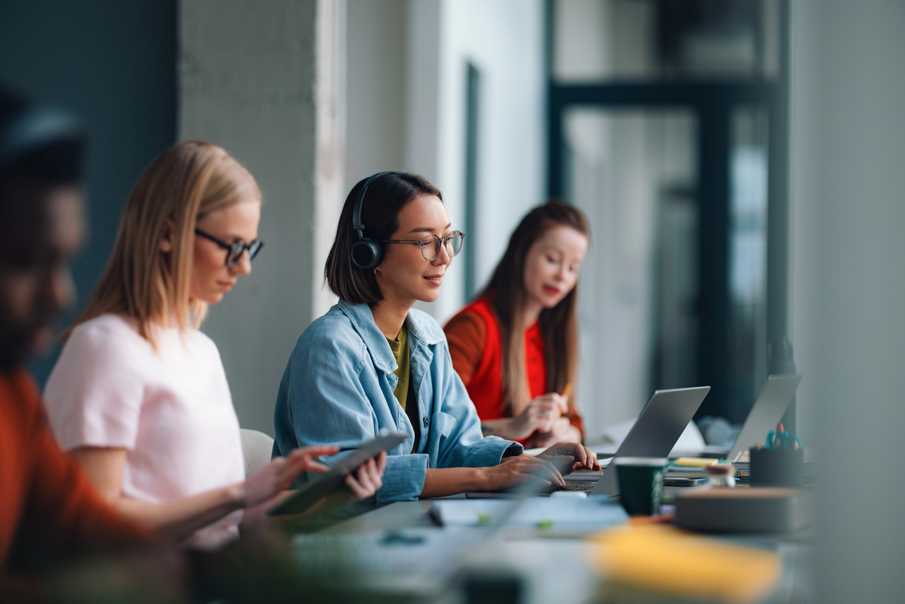 Young students collaborate in a bright university library workspace, using laptops, tablets and headphones while studying together, sharing ideas and working on a startup or class project in a modern setting.