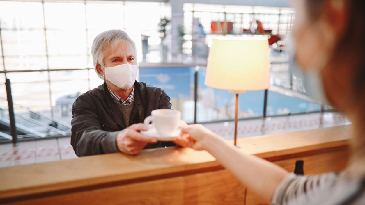 A man wearing a face mask is handing a cup of coffee to a woman at an airport.