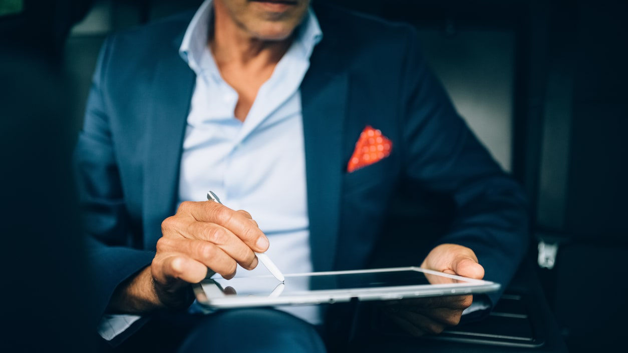 A man in a suit writing on a tablet in the back seat of a car.