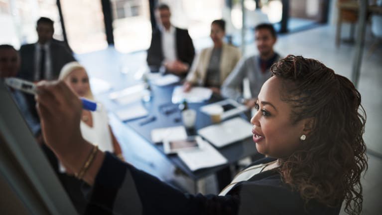 A business woman is writing on a board in front of a group of people.