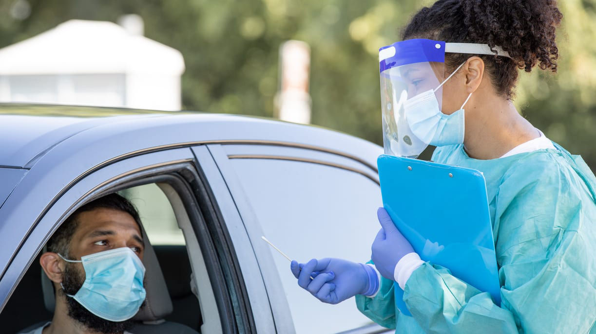 A woman wearing a surgical mask and a clipboard in front of a car.