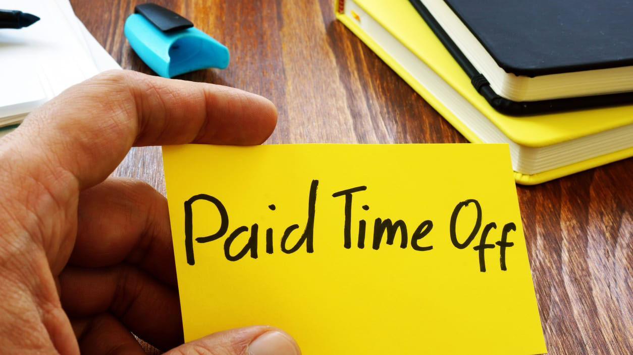 A hand holding a yellow sticky note that says paid time off.