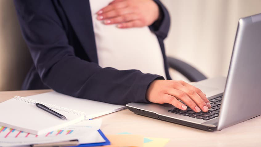 A pregnant woman working on a laptop at her desk.