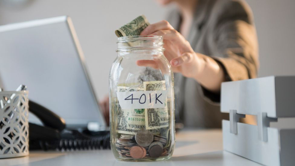 A person places cash into a jar labeled “401K” on a desk with a laptop and office supplies in the background.