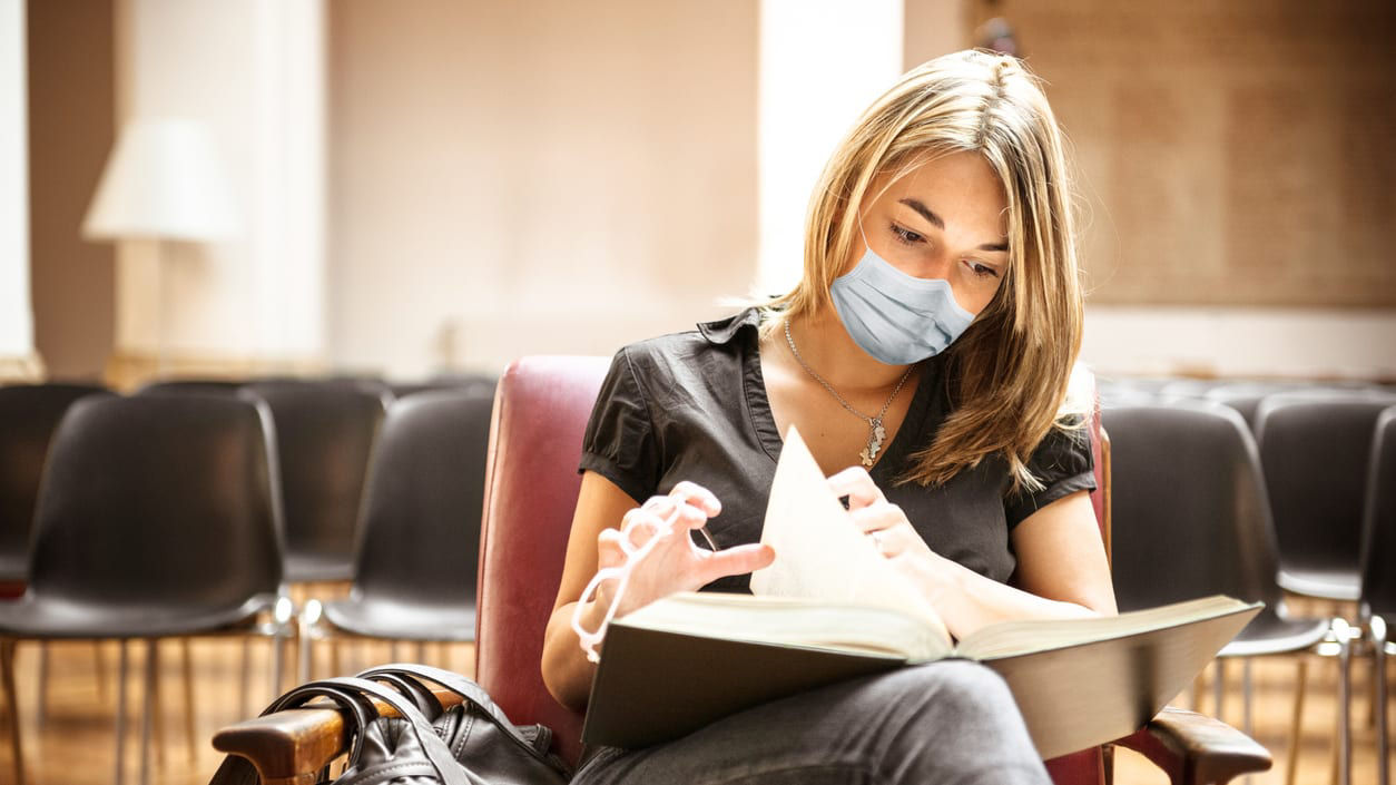A woman wearing a surgical mask sitting in a chair reading a book.