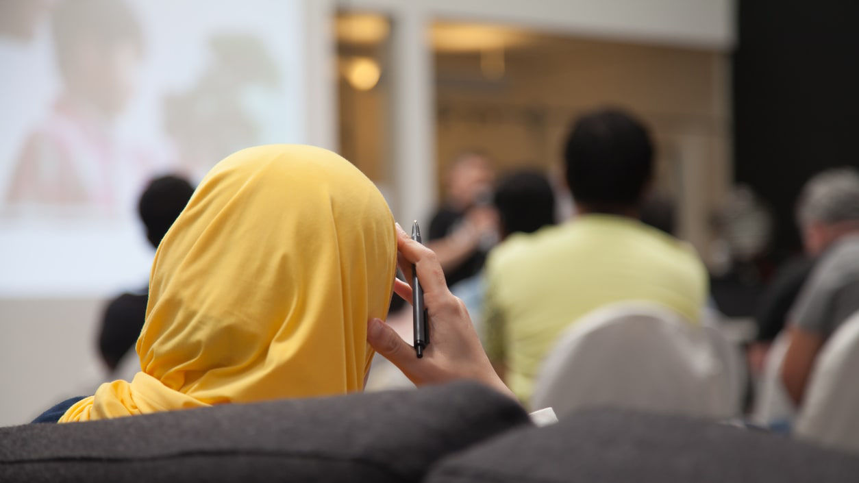 A woman wearing a yellow head scarf watching from the audience at a conference.