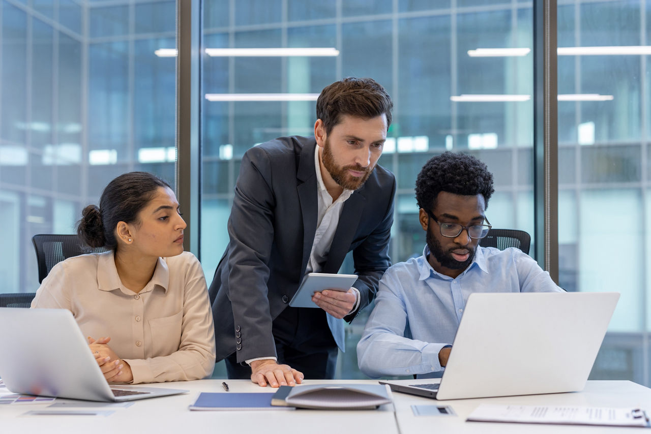 A diverse group of professional colleagues working together in an office space. They are focused and engaged, using a laptop and a tablet. This image highlights teamwork and collaboration.