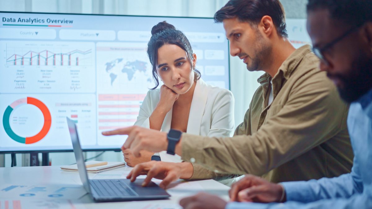 Three employees in professional dress, including two men and a woman, look at laptops and analyze the business value of a program.