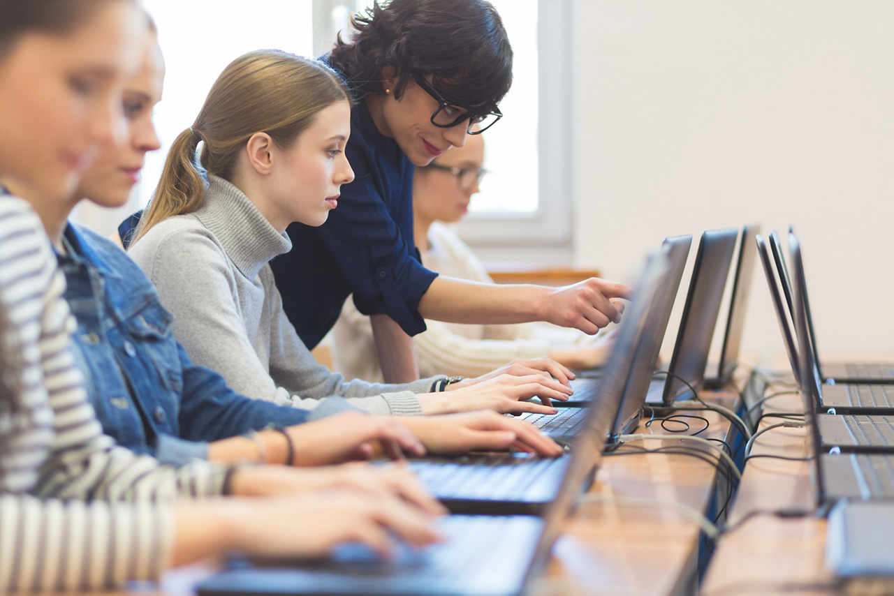 Female students learning computer programming with teacher. Multiracial group of girls in computer lab coding on laptops.