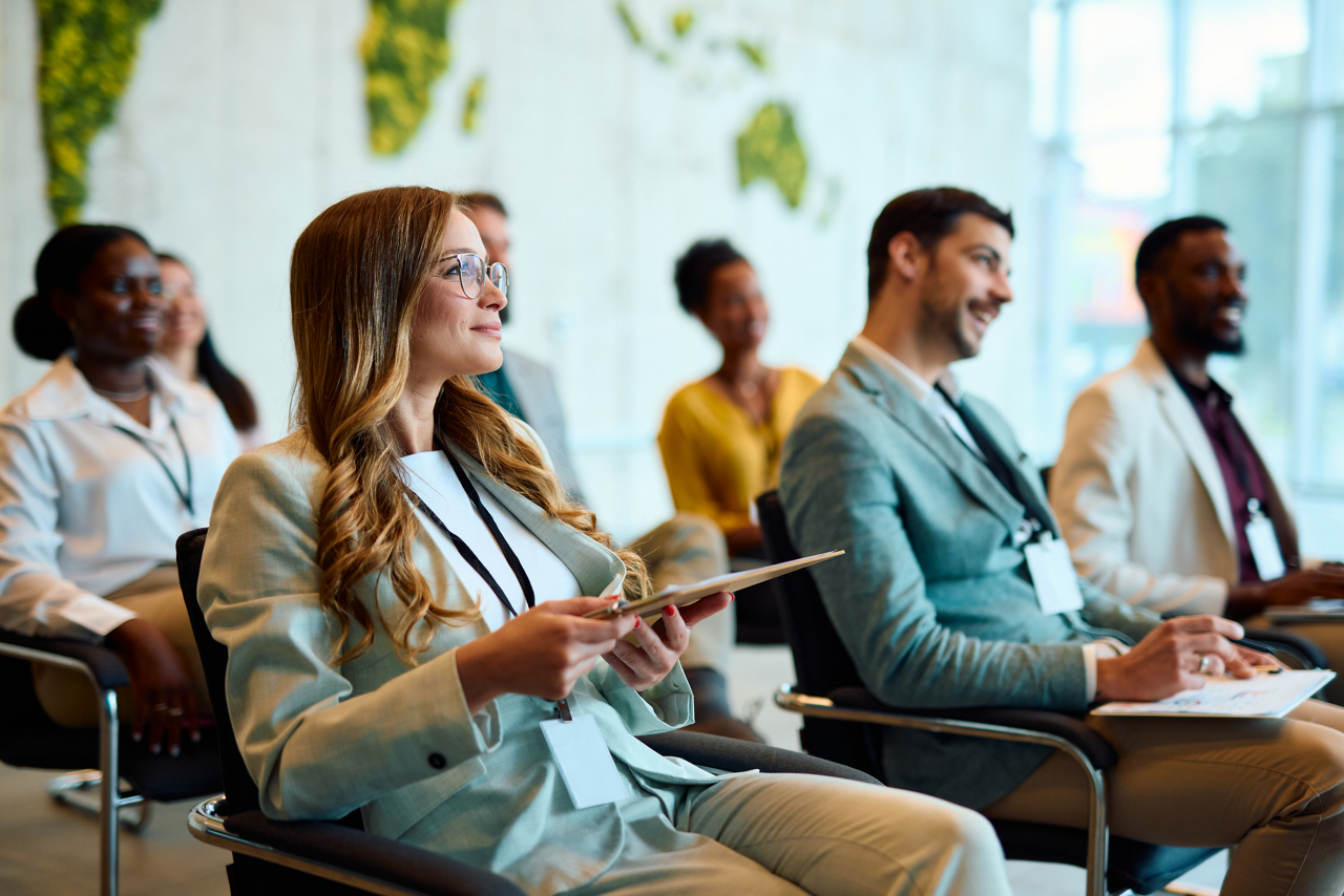 Group of diverse businesspeople seated in chairs, attentively listening to a presentation during a business convention or seminar, engaged in learning and professional development