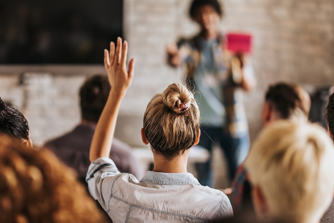 Rear view of casual businesswoman raising her hand to ask the question on education event in a board room.