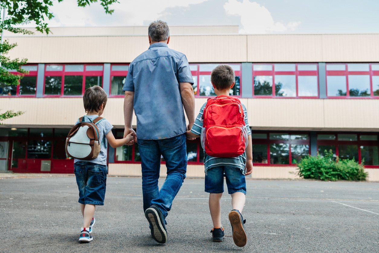 Image of a dad holding the hands of two boys wearing backpacks walking toward a school