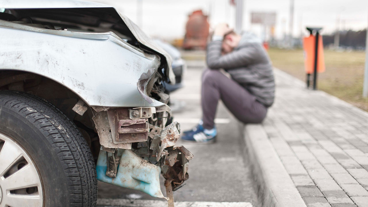A woman sits next to a car that has been damaged in an accident.
