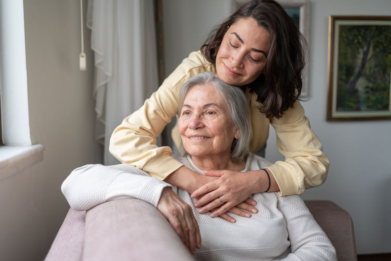  A woman gently hugs her senior mother who is sitting down.