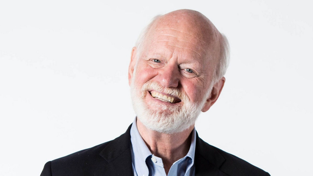 An older man smiling with a white beard and blue dress shirt.