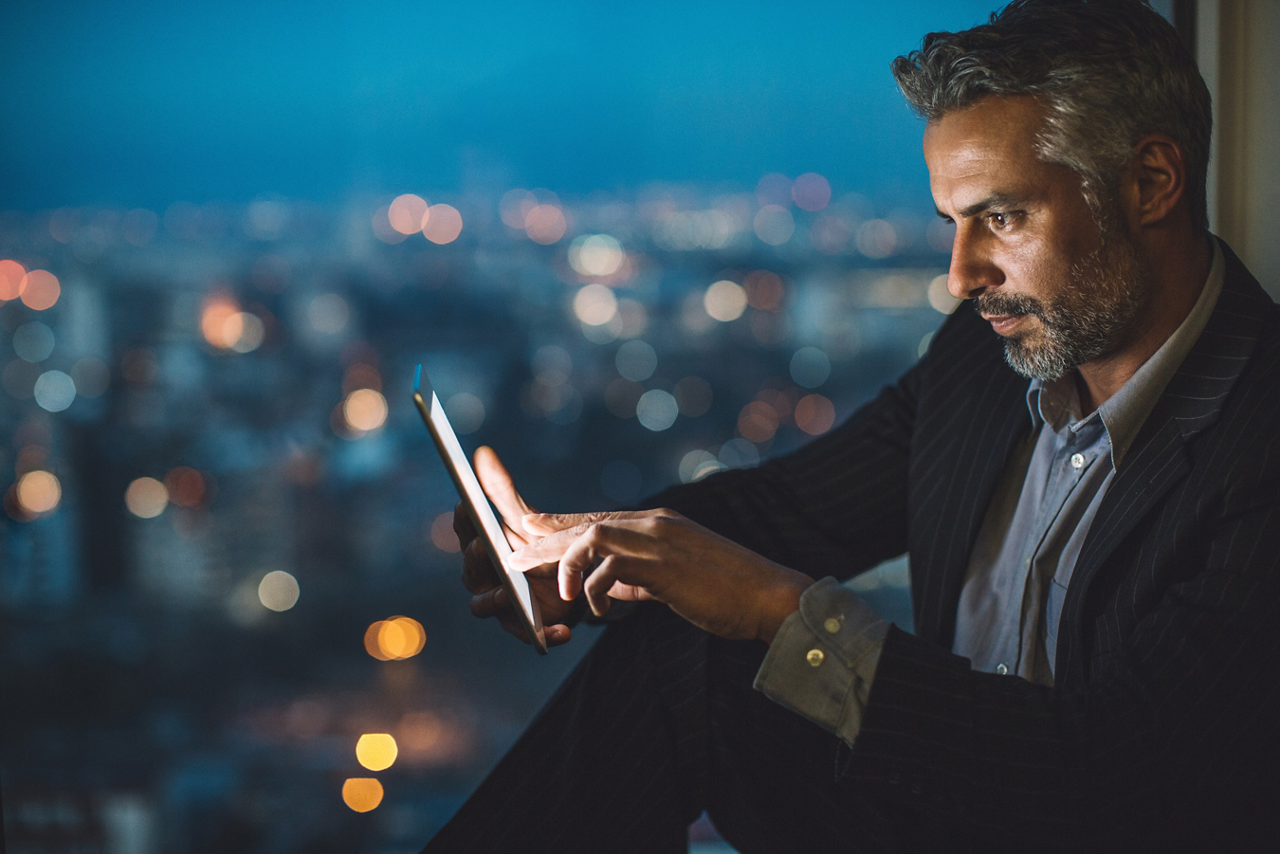Businessman sitting on a windowsill at night and looking at a digital tablet, with cityscape at the background and copy space.