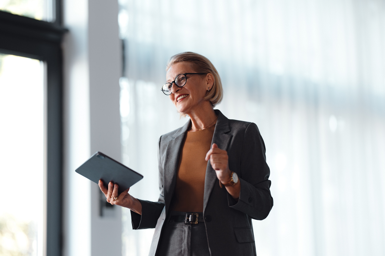 Portrait of a beautiful senior female presenter dressed in business clothes. She is standing in the office or a meeting room, holding a digital tablet. She is smiling and looking away.