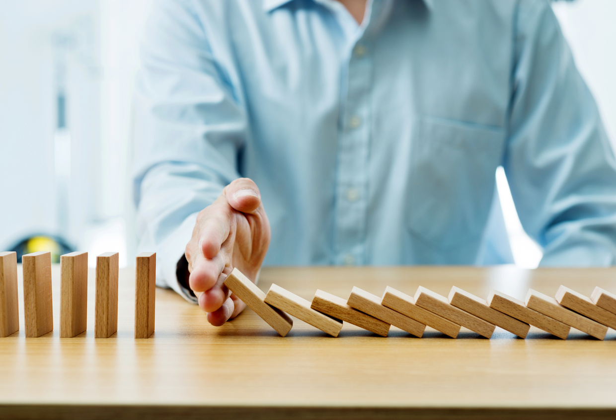 A hand in a light blue shirt stopping a line of falling wooden dominoes on a wooden surface, symbolizing intervention or prevention.