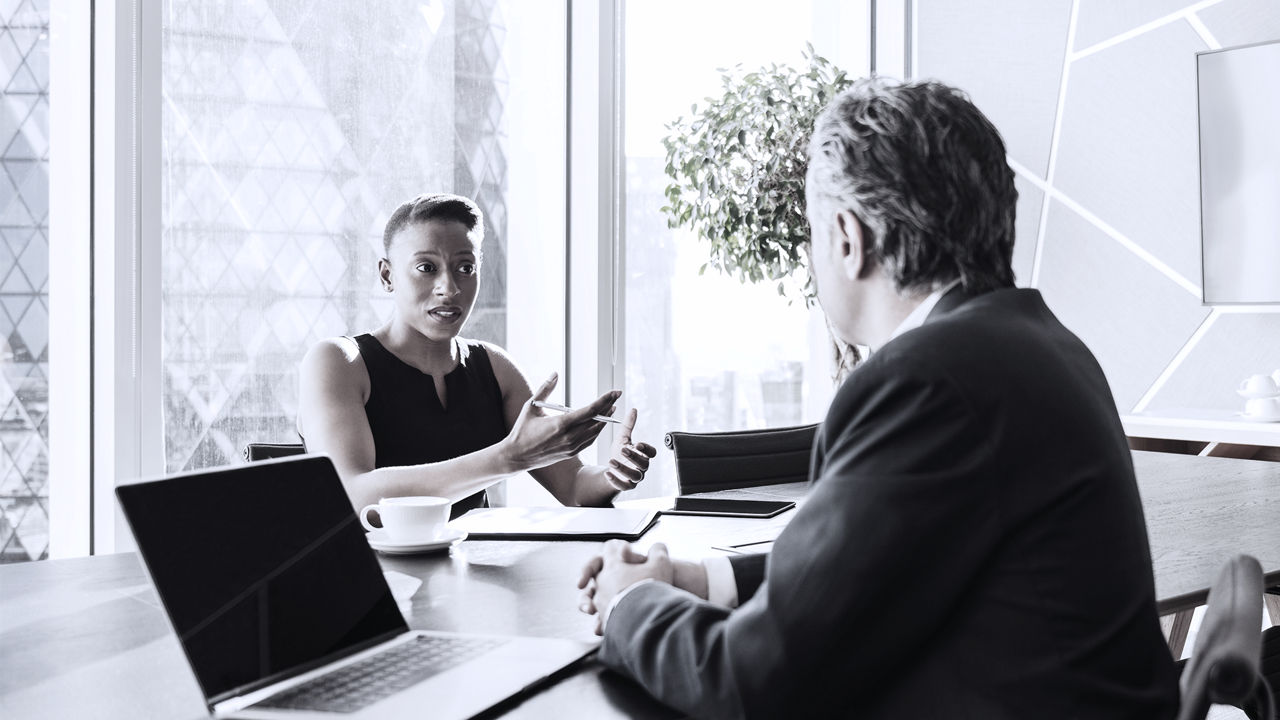 Female executive on left talks to a male executive on right, both sitting across from each on other in office at a table