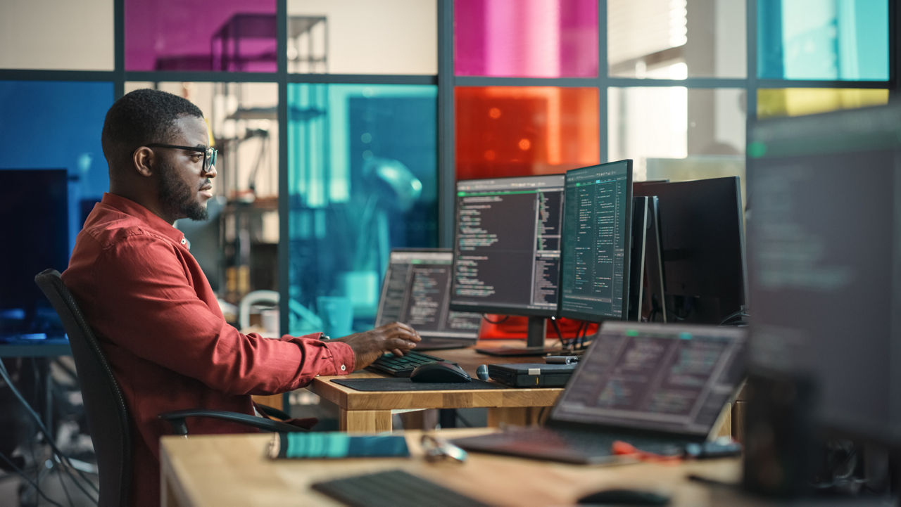 A black man writes lines of code in a room filled with computer screens.
