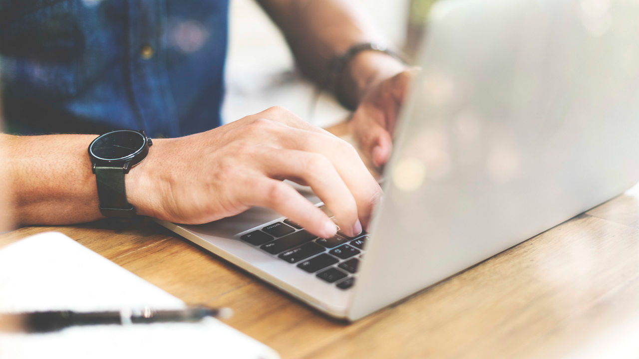 A man typing on a laptop in front of a wooden table.