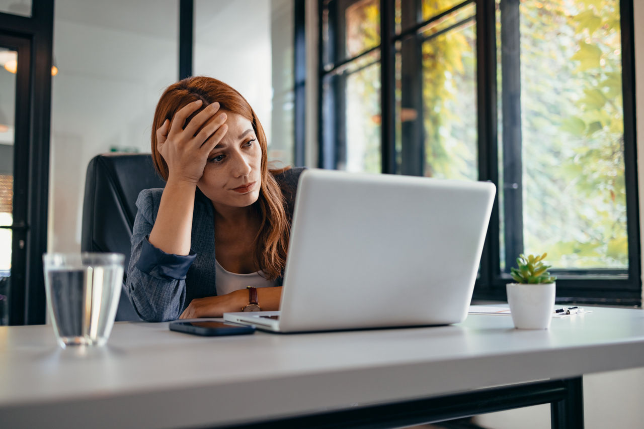 A woman is sitting at a desk in an office with a laptop and a glass of water, seemingly feeling stressed and overwhelmed by her work.