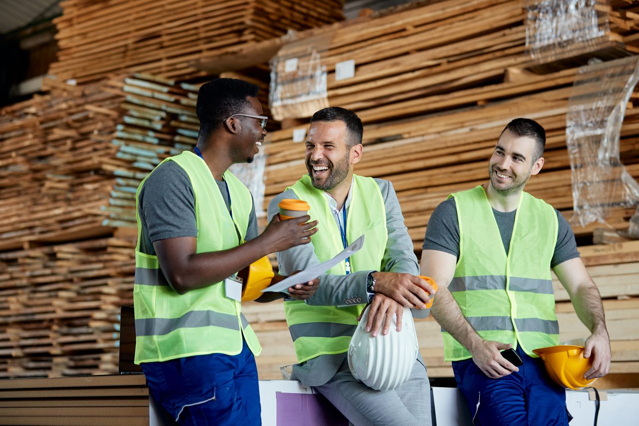 Happy workers having fun while talking with their manager on coffee break at lumber distribution warehouse.