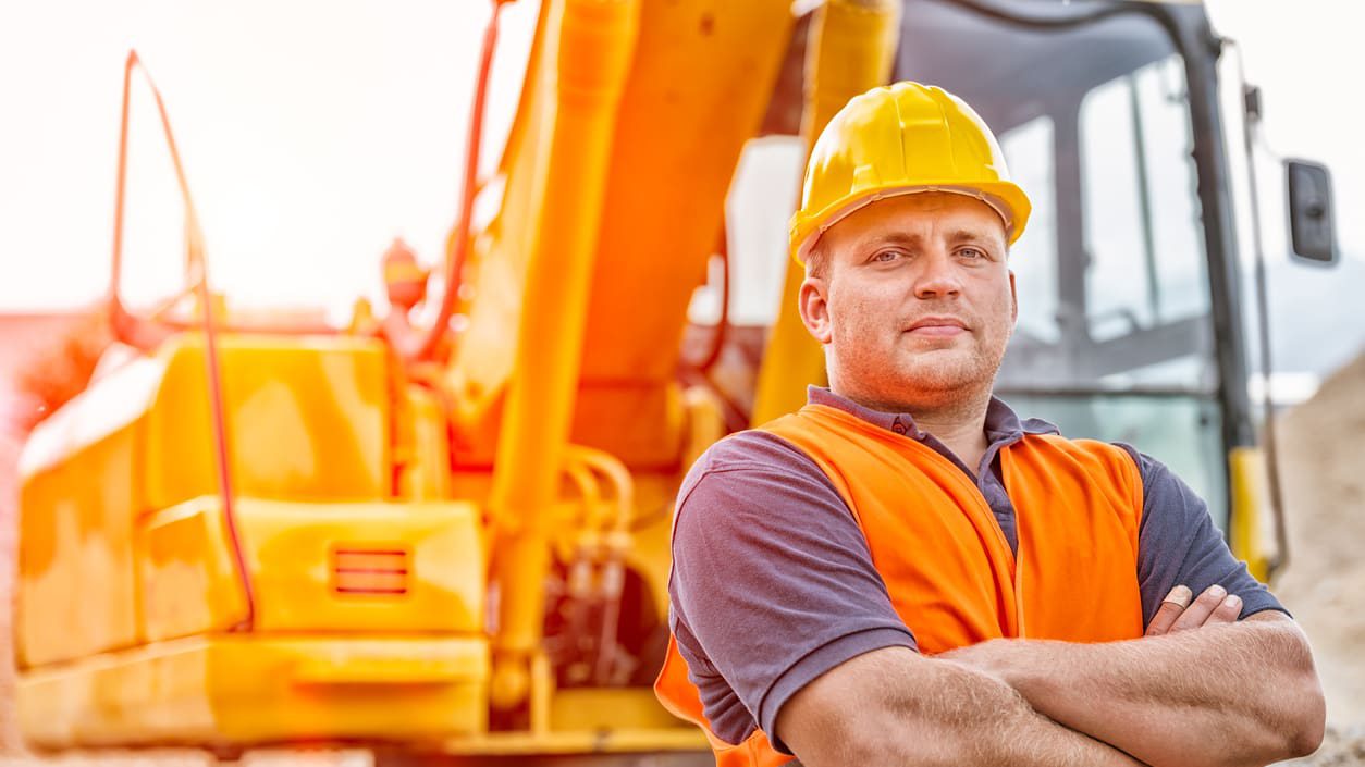 A construction worker wearing a hard hat, arms crossed, with a building crane behind him.